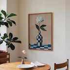 a dining area with a wooden table, a vase with a single pink flower, and a framed painting of a white flower in a blue and white checkered vase hanging on the wall.