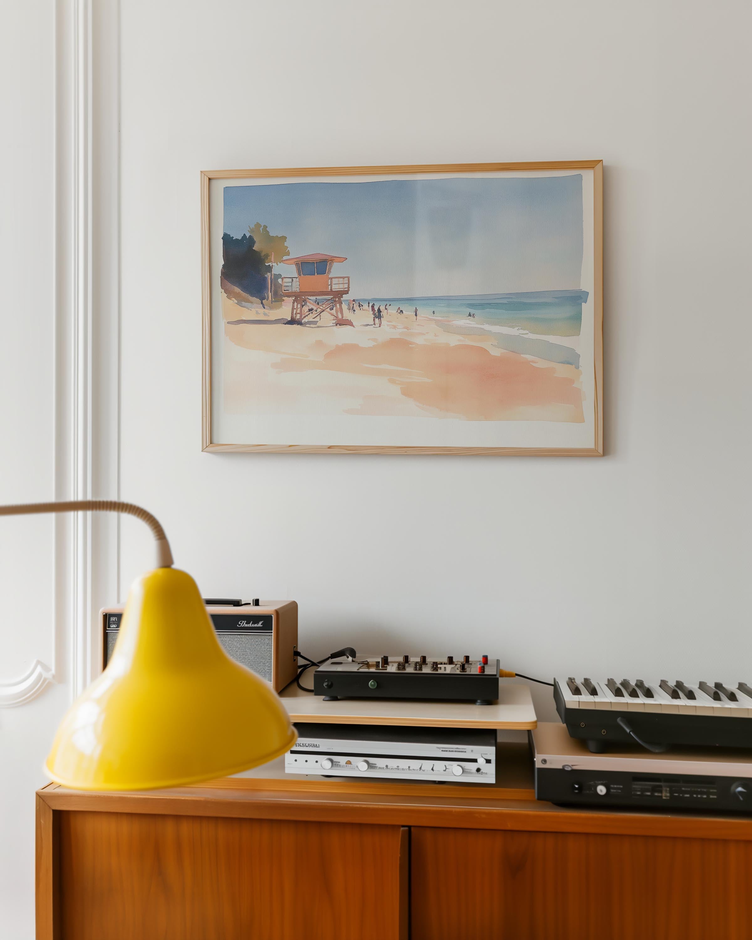 a framed painting of a beach scene hanging on a wall above a wooden cabinet with various electronic equipment and a yellow lamp on top.