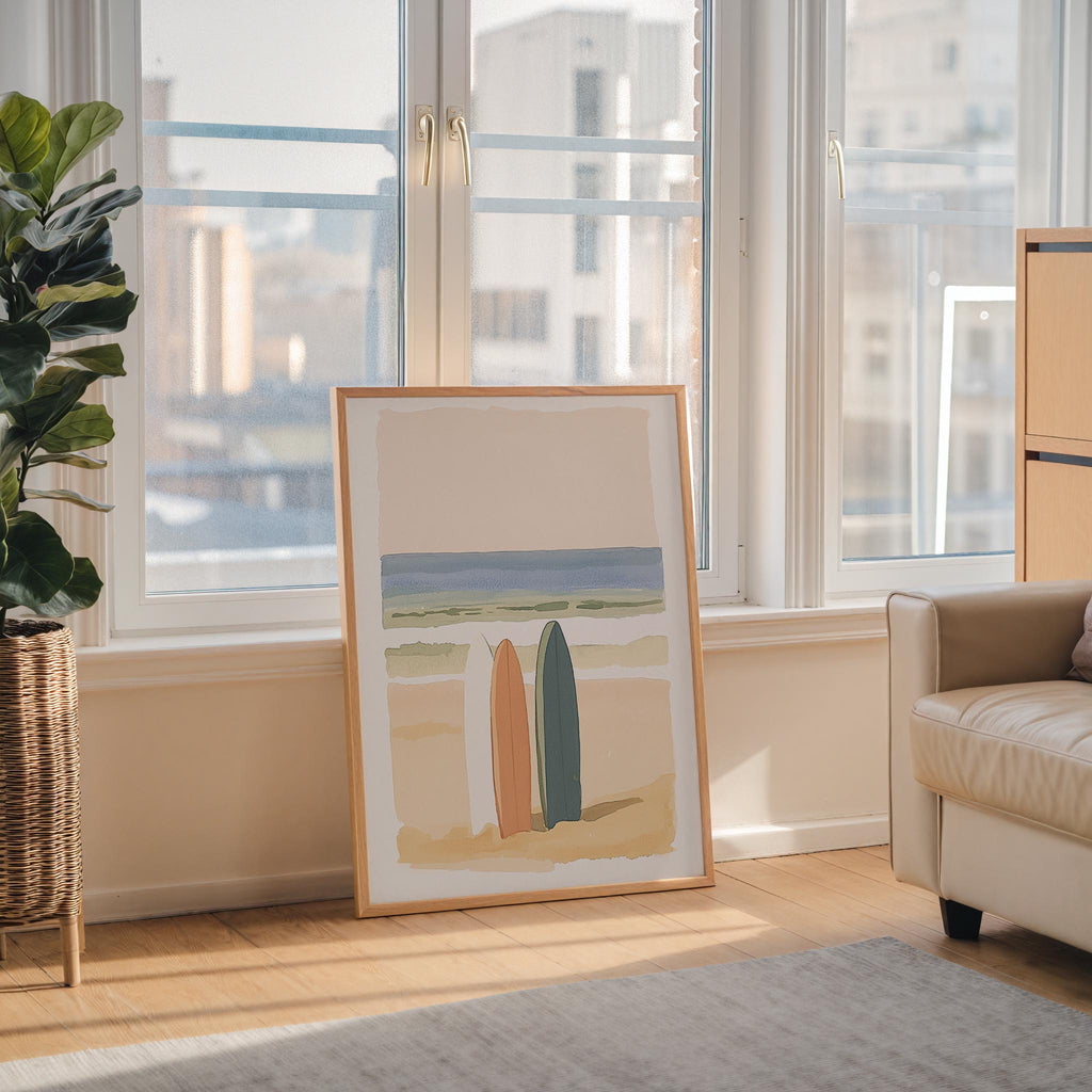 a large framed artwork depicting a beach scene with two surfboards, placed on a wooden floor in a living room with large windows overlooking a cityscape.