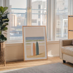 a large framed artwork depicting a beach scene with two surfboards, placed on a wooden floor in a living room with large windows overlooking a cityscape.