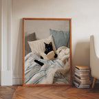 A framed illustration of a black cat lying on a bed and reading a book.
