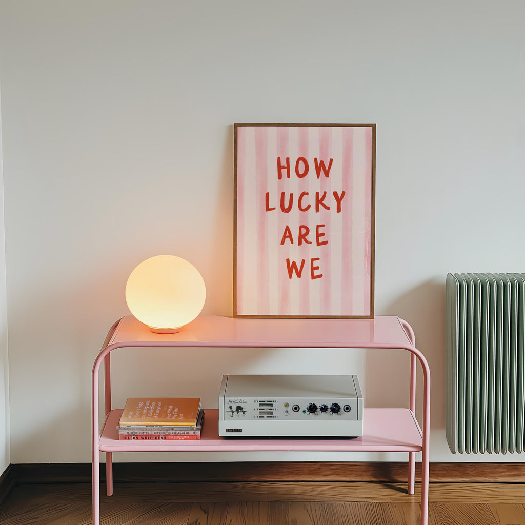 a pink metal table with a lamp, a framed poster with the text "HOW LUCKY ARE WE", and a small white speaker on the table.
