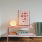 a pink metal table with a lamp, a framed poster with the text "HOW LUCKY ARE WE", and a small white speaker on the table.