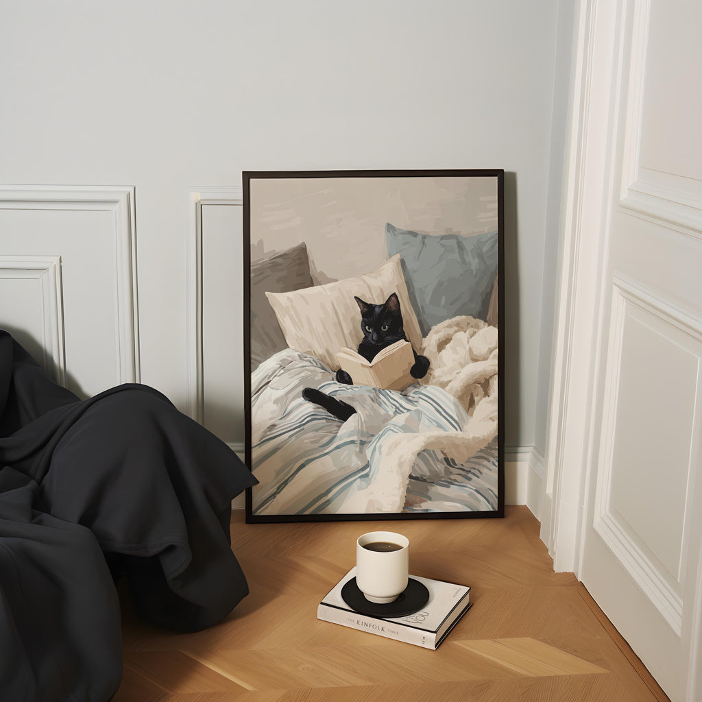 A framed painting of a black cat lying on a bed, with a cup of coffee and a book on the floor in front of it.