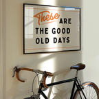 a framed sign with the text "These are the good old days" hanging on a wall next to a bicycle.