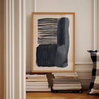 a framed abstract artwork with bold black and white stripes, displayed on a white wall next to a stack of books.