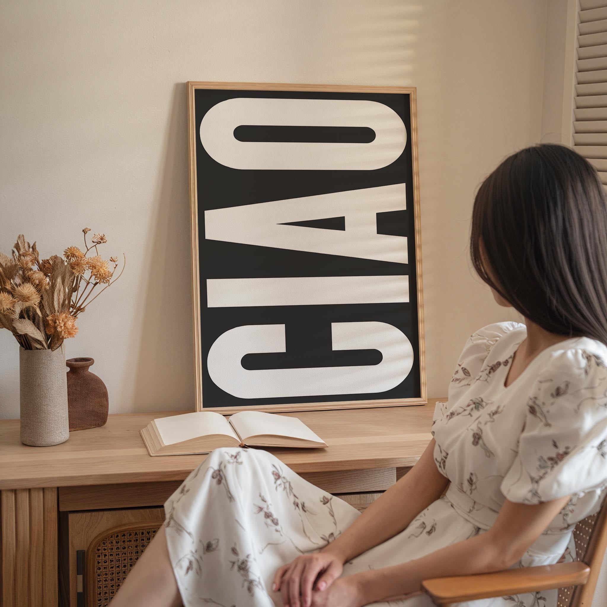 A woman in a white dress is sitting at a desk with an open book and a vase of flowers, with a large black and white sign that says "Ciao" in the background.