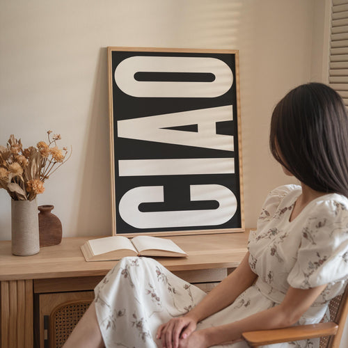 A woman in a white dress is sitting at a desk with an open book and a vase of flowers, with a large black and white sign that says "Ciao" in the background.