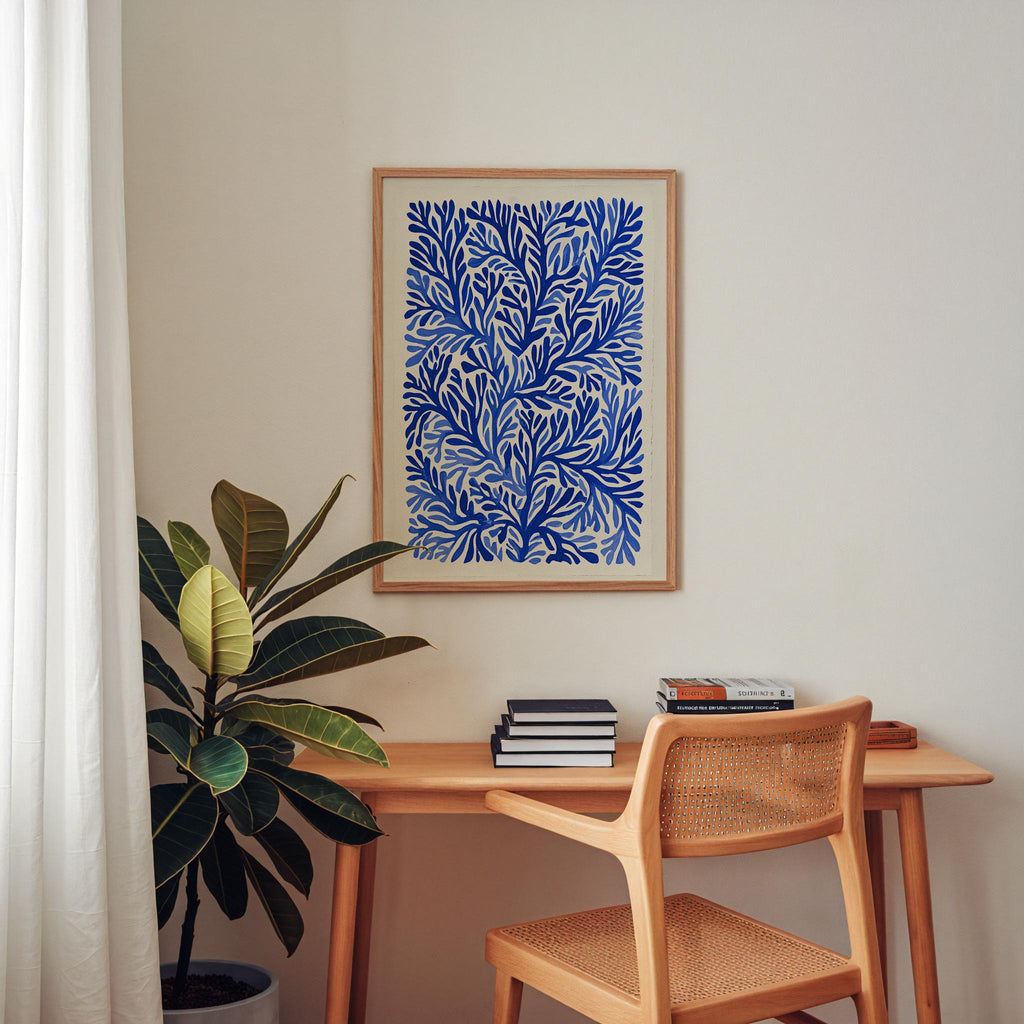 a cozy corner of a room with a wooden desk, a woven chair, and a potted plant. A framed blue and white abstract painting hangs on the wall above the desk, which is adorned with several books and a stack of magazines.