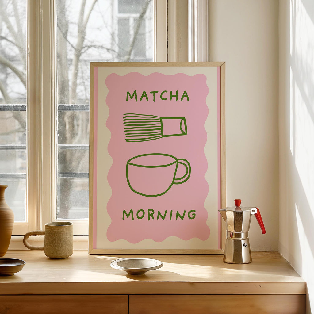 a framed poster with a pink background and a green illustration of a cup and a whisk, along with the text "MATCHA MORNING". The poster is placed on a wooden shelf, and there is a coffee maker and a ceramic mug on the shelf as well.