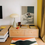 a cozy corner of a room with a wooden dresser, a lamp, a framed painting of a dog reading a book, and a stack of books.