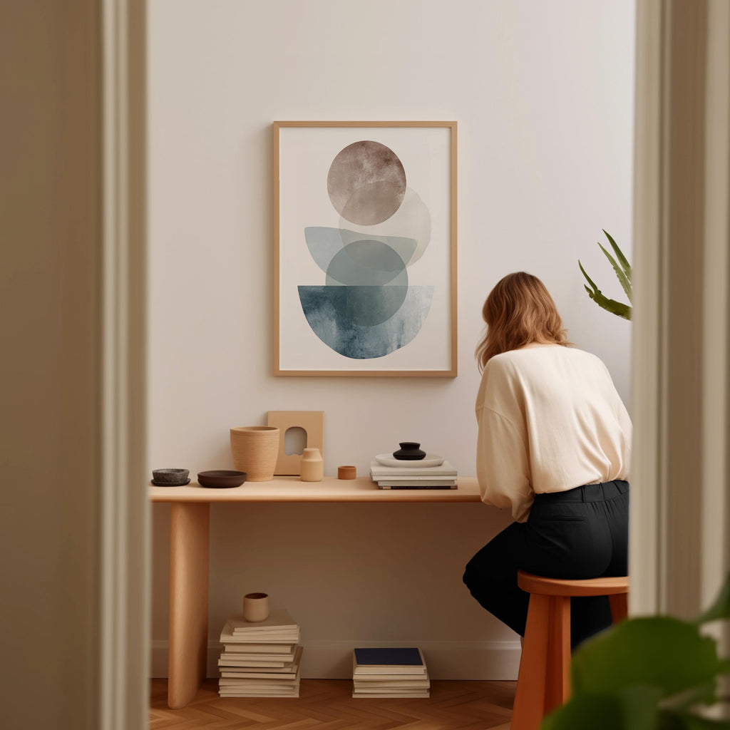 A woman is sitting at a wooden desk, surrounded by various objects, including a framed abstract art piece, a stack of books, and a plant.