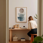 A woman is sitting at a wooden desk, surrounded by various objects, including a framed abstract art piece, a stack of books, and a plant.