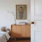 a cozy bedroom scene with a wooden dresser, a framed artwork of a woman reading, and a bed with a gray comforter.
