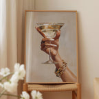 A framed painting of a hand holding a martini glass, with a bracelet on the wrist.
