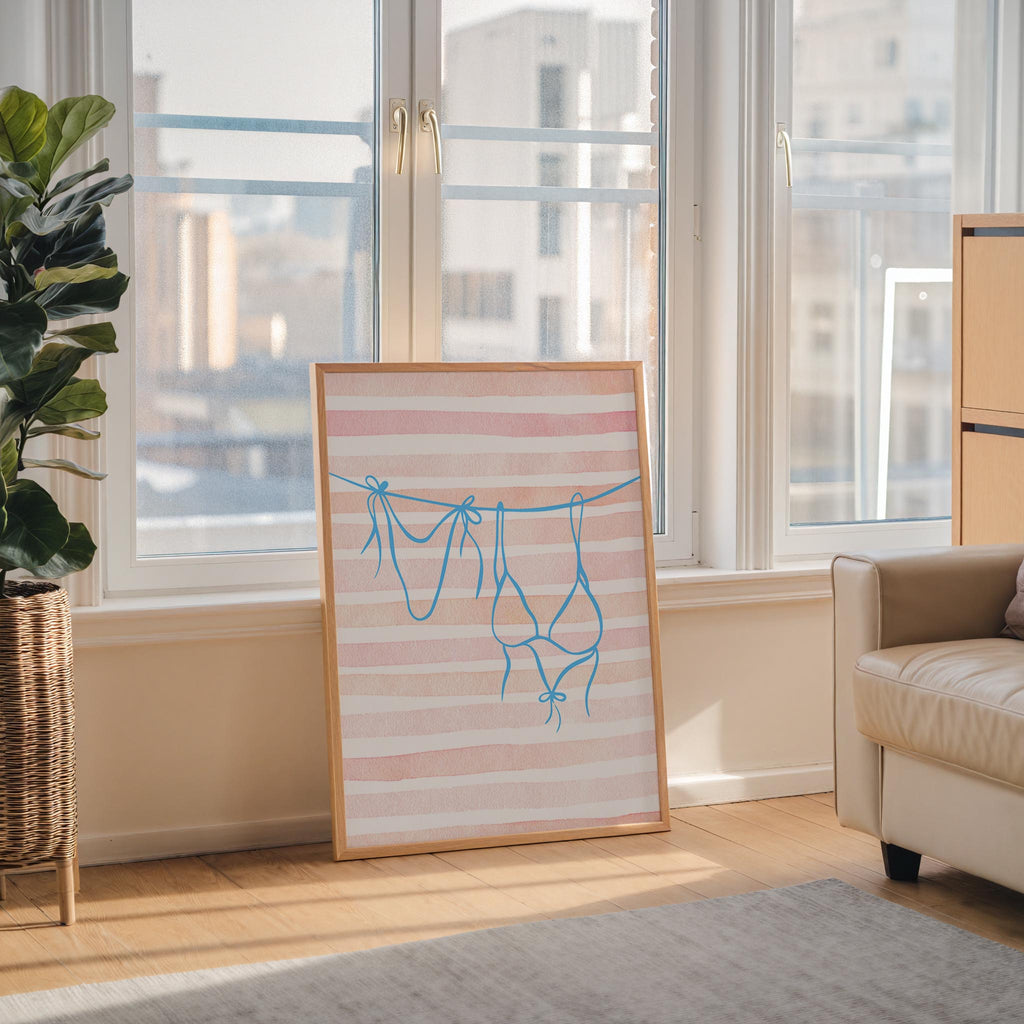 a framed artwork depicting a pair of blue swimsuits hanging on a clothesline, placed on a wooden floor in a room with large windows overlooking a cityscape.