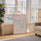 a framed artwork depicting a pair of blue swimsuits hanging on a clothesline, placed on a wooden floor in a room with large windows overlooking a cityscape.