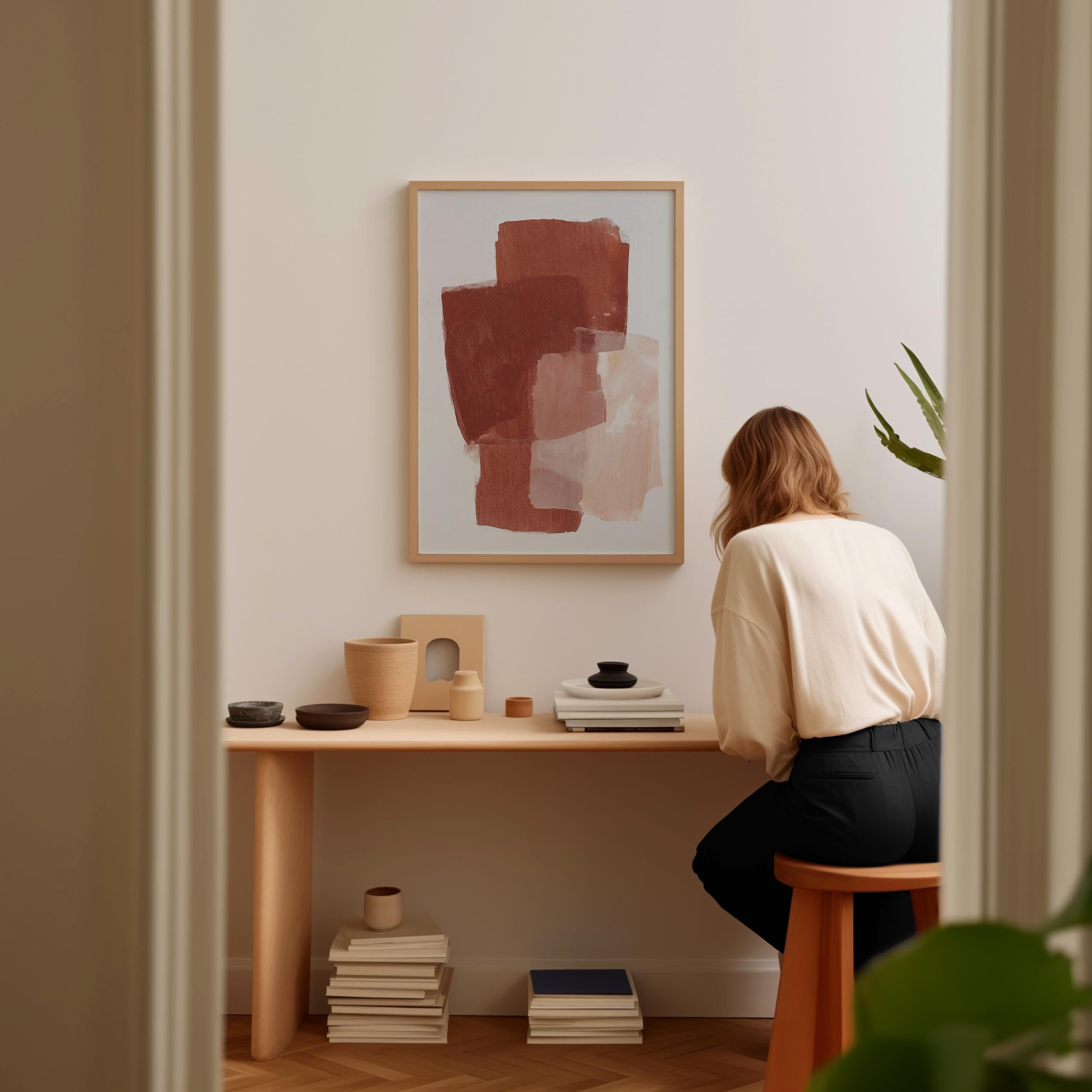 A woman is sitting at a wooden desk, surrounded by various objects, including a framed abstract painting, a stack of books, and a plant.