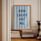 a framed poster with the text "HOW LUCKY ARE WE" displayed on it, placed on a shelf next to a stack of books.