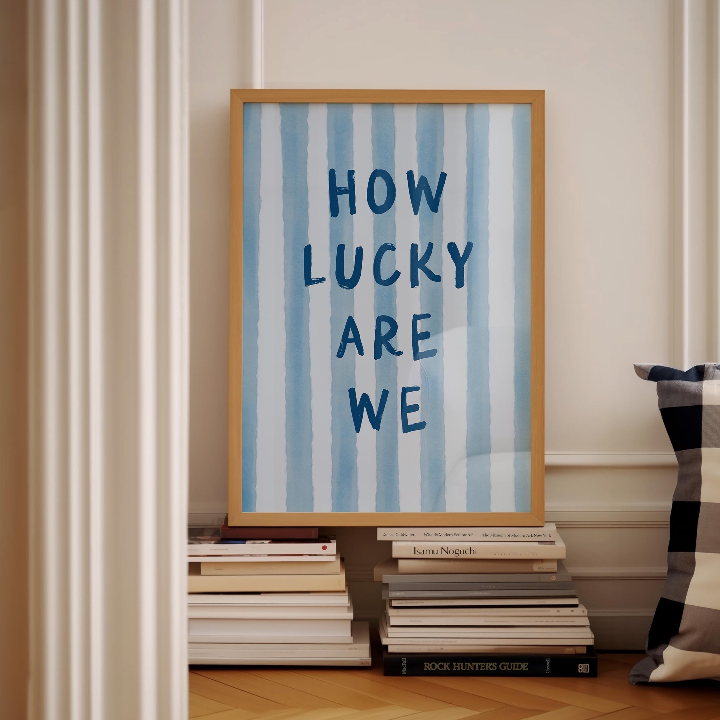 a framed poster with the text "HOW LUCKY ARE WE" displayed on it, placed on a shelf next to a stack of books.