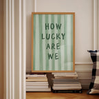 a framed poster with the text "HOW LUCKY ARE WE" displayed on it, placed on a shelf next to a stack of books.