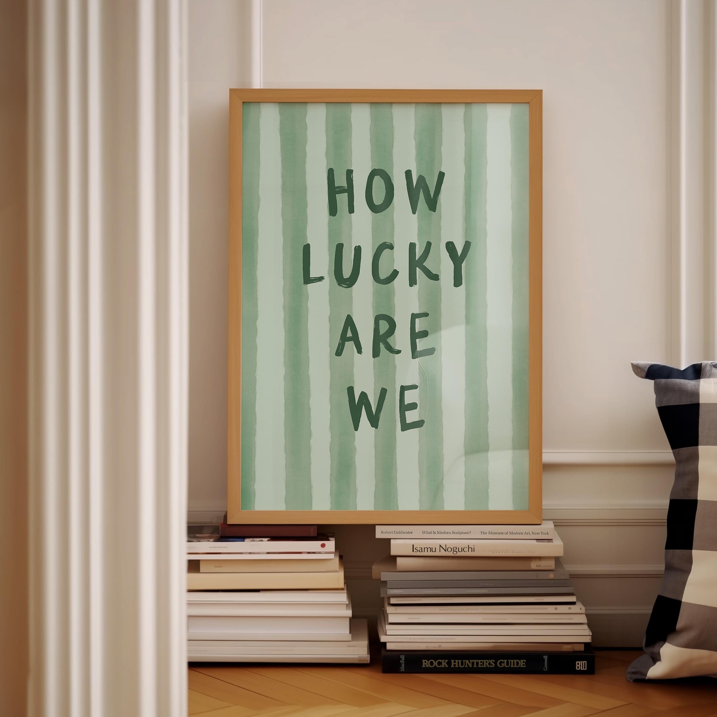a framed poster with the text "HOW LUCKY ARE WE" displayed on it, placed on a shelf next to a stack of books.