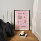 a framed poster with the text "MATCHA MORNING" and illustrations of a cup and a whisk, placed on a wooden floor next to a book and a cup of coffee.