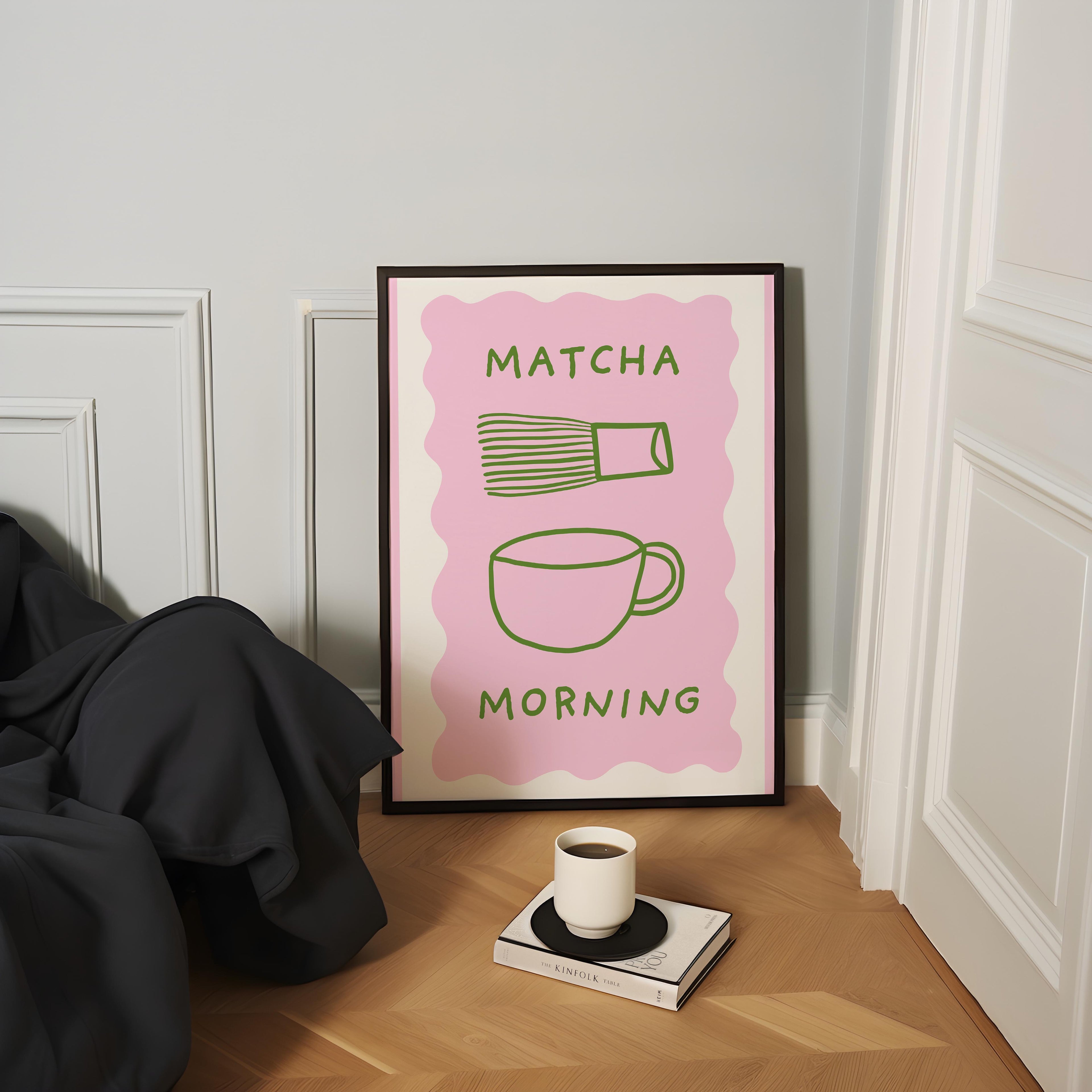 a framed poster with the text "MATCHA MORNING" and illustrations of a cup and a whisk, placed on a wooden floor next to a book and a cup of coffee.