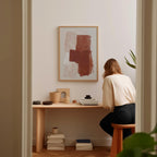 A woman is sitting at a wooden desk, surrounded by various objects, including a framed abstract painting, a stack of books, and a plant.