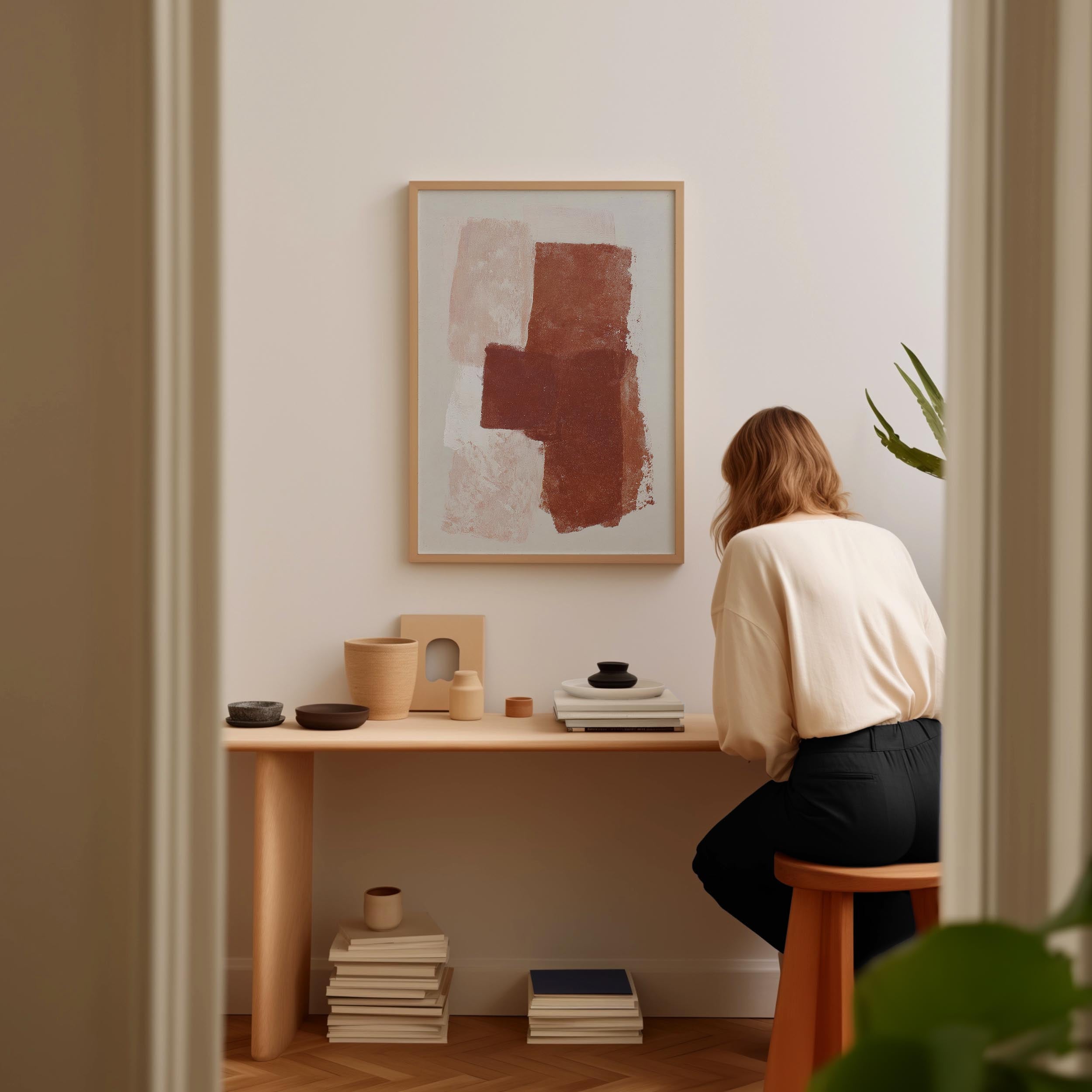 A woman is sitting at a wooden desk, surrounded by various objects, including a framed abstract painting, a stack of books, and a plant.