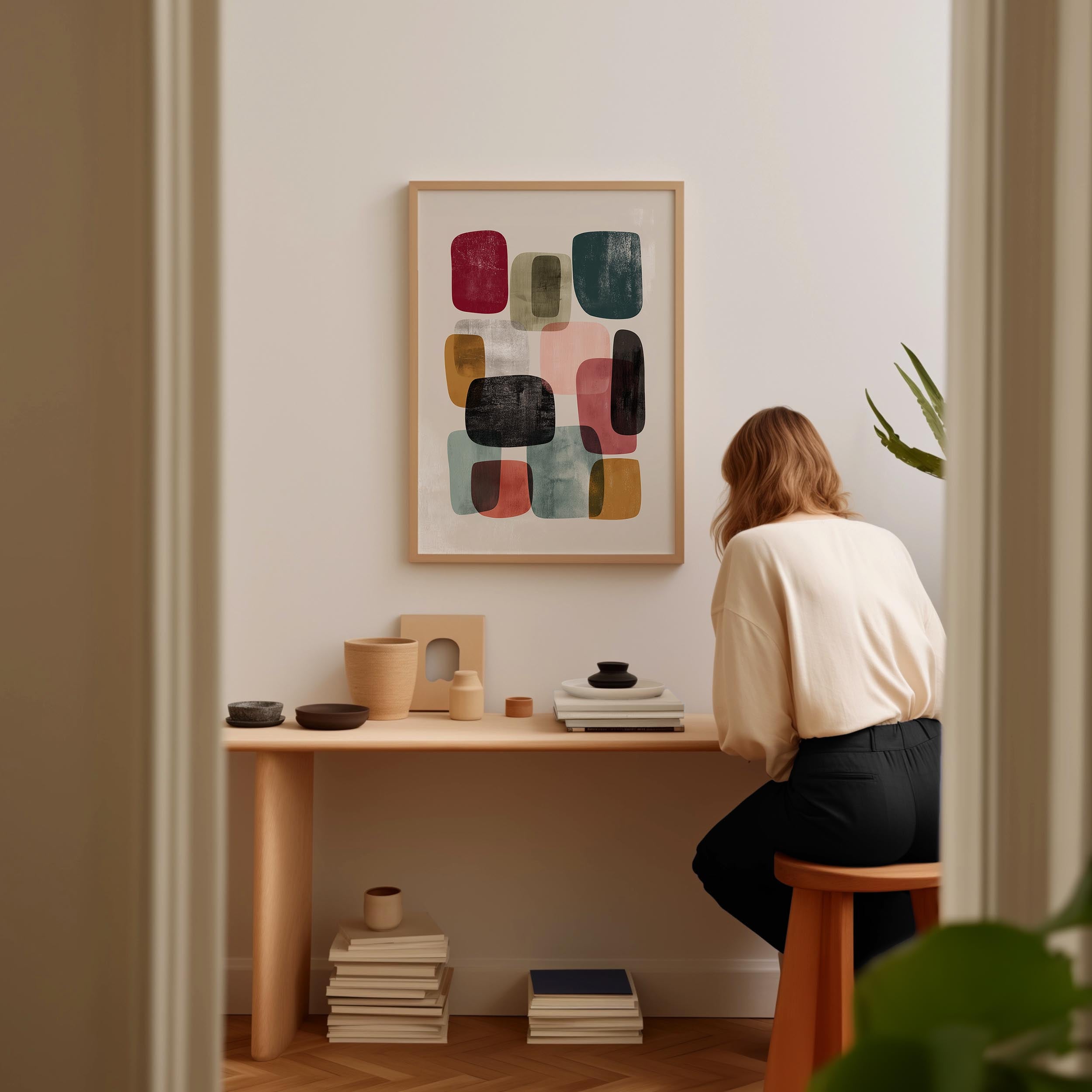 A woman is sitting at a wooden desk, surrounded by various objects, including a stack of books, a plant, and a framed abstract art piece on the wall.