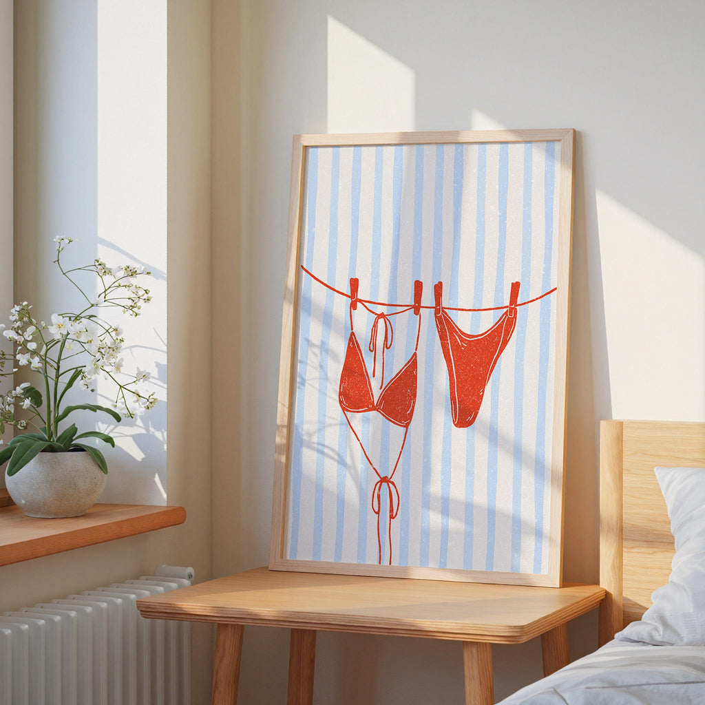 a framed artwork depicting two red bikini bottoms hanging on a clothesline, with a white and blue striped background.