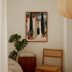a cozy corner of a room with a painting of a house, a potted plant, and a wooden chair.
