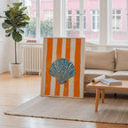 a living room with a large framed artwork of a seashell on an orange and white striped background, placed on a rug in front of a window. The room also includes a sofa, a coffee table, and a potted plant.
