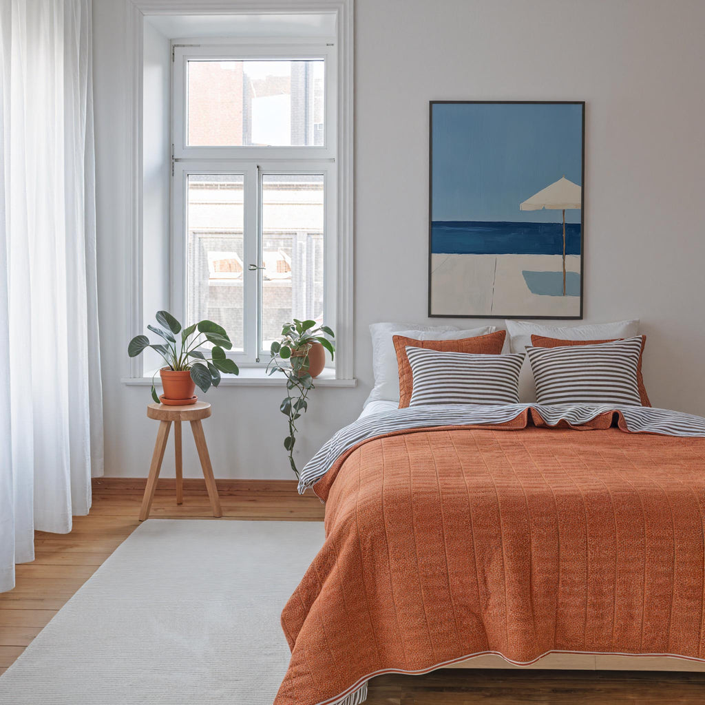 a cozy bedroom with a large bed, a wooden stool with a potted plant, and a framed artwork of a beach scene on the wall.