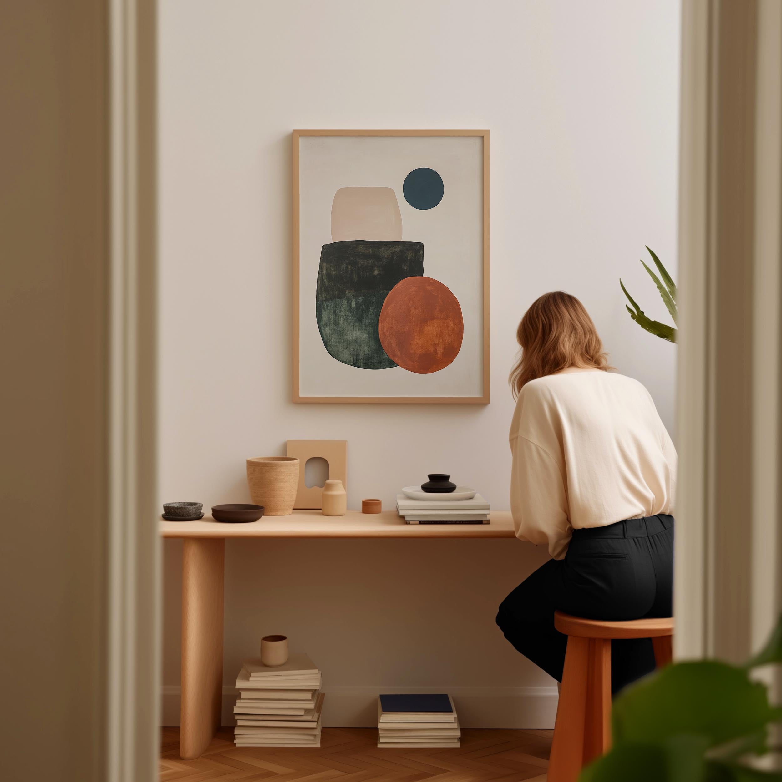 A woman is sitting at a wooden desk, surrounded by various objects, including a framed abstract artwork, a stack of books, and a plant.