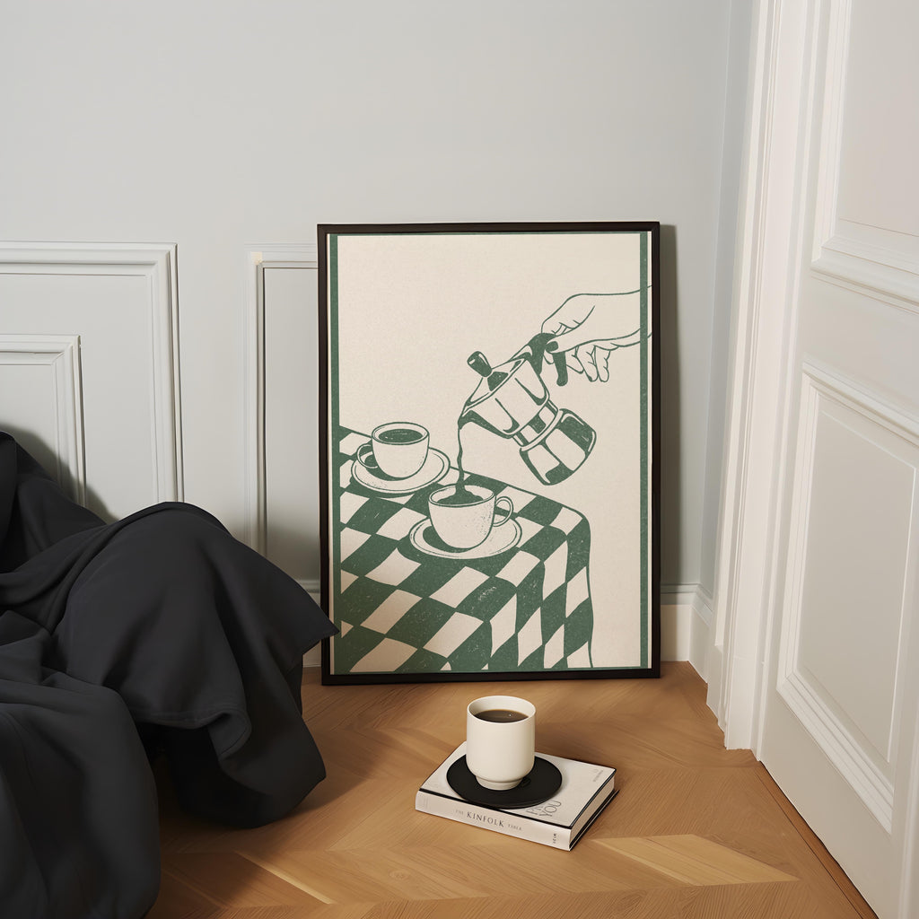 a cozy corner with a framed illustration of a hand pouring coffee into two cups, a white mug, and a book on a wooden floor.