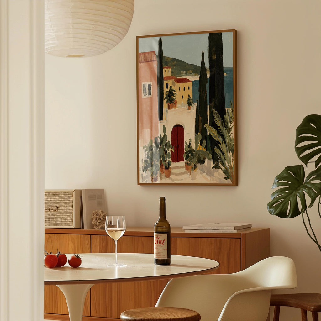 a dining area with a table set for two, a bottle of wine, and a framed painting of a coastal town.