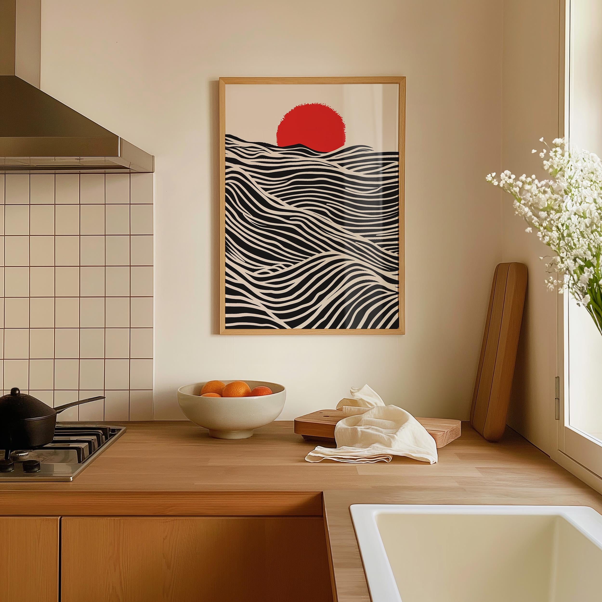 a kitchen counter with a bowl of fruit, a cutting board, and a vase of flowers. Above the counter, there is a framed artwork depicting a black and white wave with a red sun in the center.