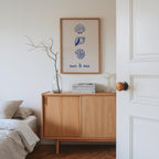 a bedroom with a wooden dresser, a framed poster of seashells, a vase with branches, and a stack of books.