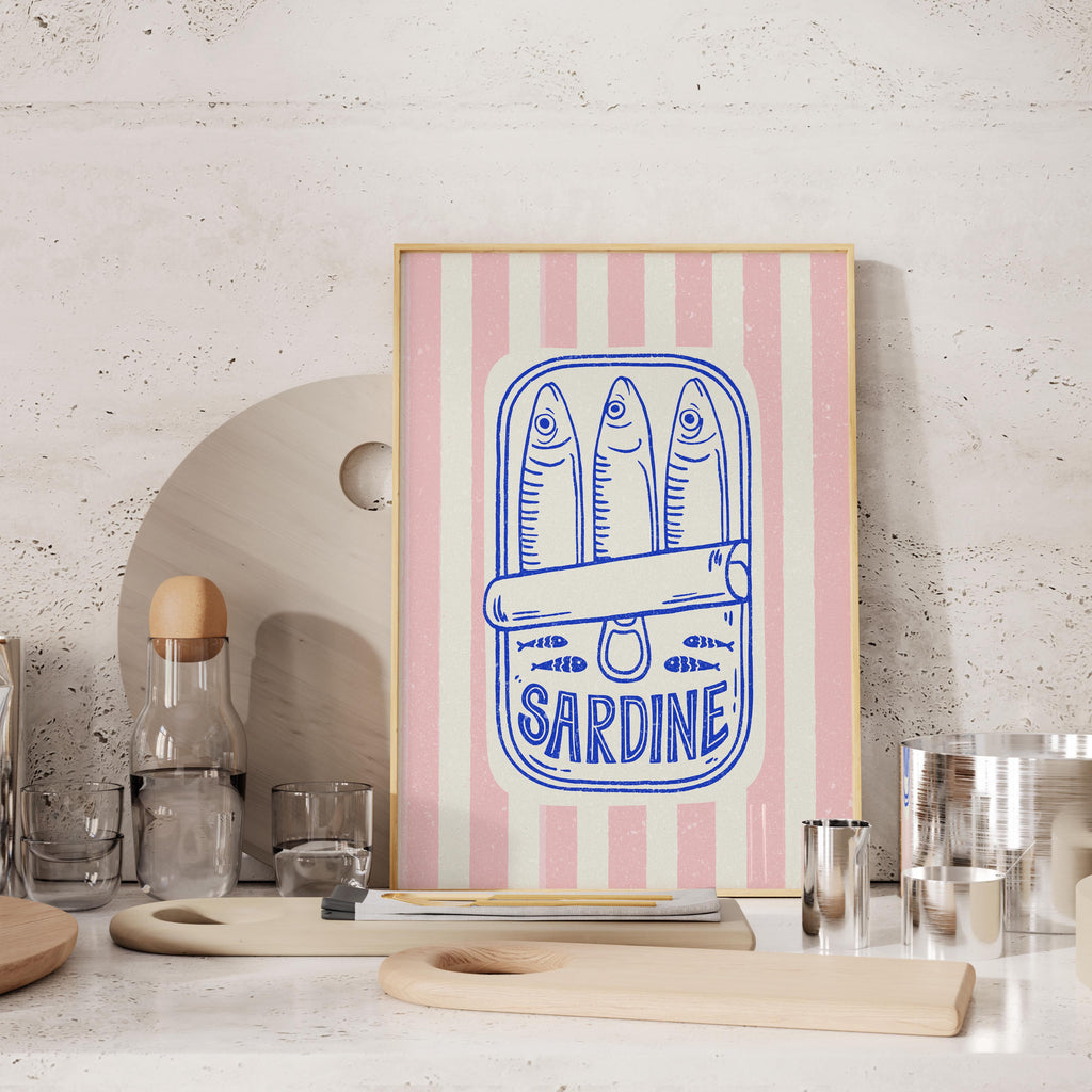 a framed artwork with a pink and white striped background, depicting a can of Sardine with three fish. The artwork is placed on a table with various kitchen items, such as a bottle, a glass, and a wooden cutting board.