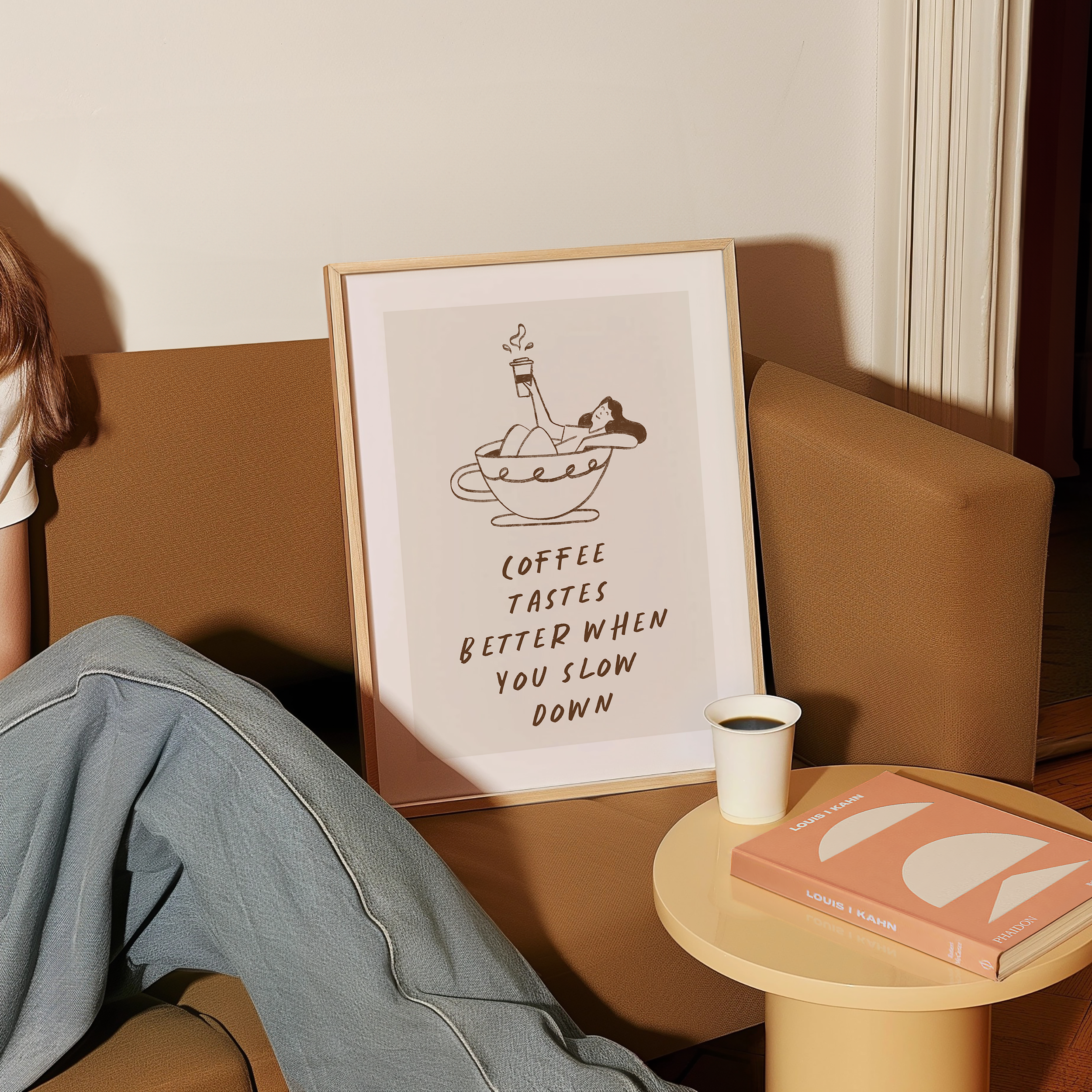 A framed poster with a coffee cup illustration and text is displayed on a brown couch, accompanied by a person's legs and a small table with a book and a cup of coffee.