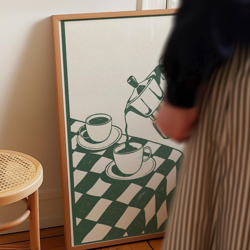 a person standing next to a framed picture of two cups of coffee on a checkered tablecloth.