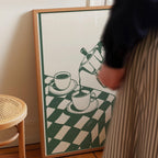 a person standing next to a framed picture of two cups of coffee on a checkered tablecloth.