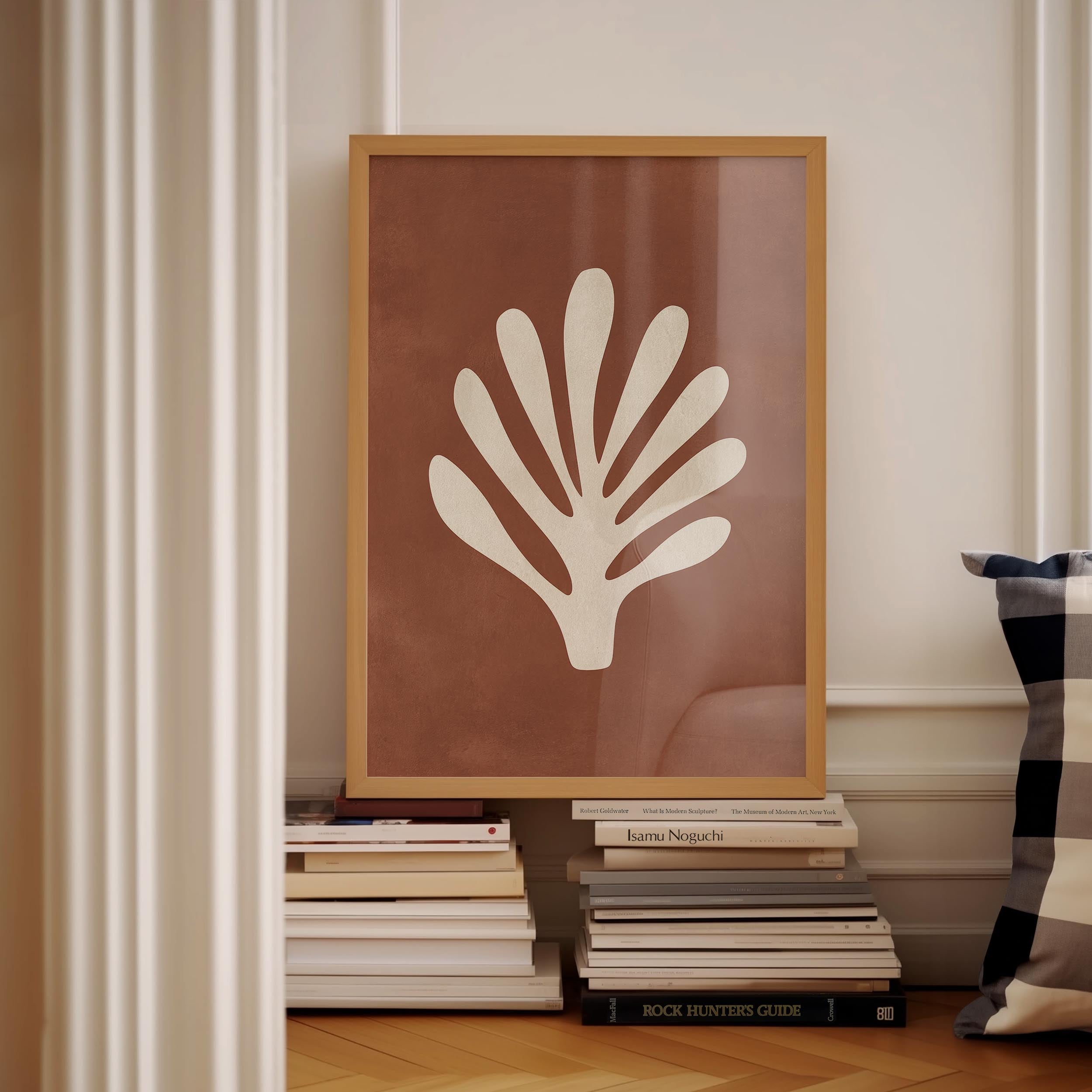 a framed artwork of a white leaf-like design on a brown background, placed on a shelf among a stack of books.