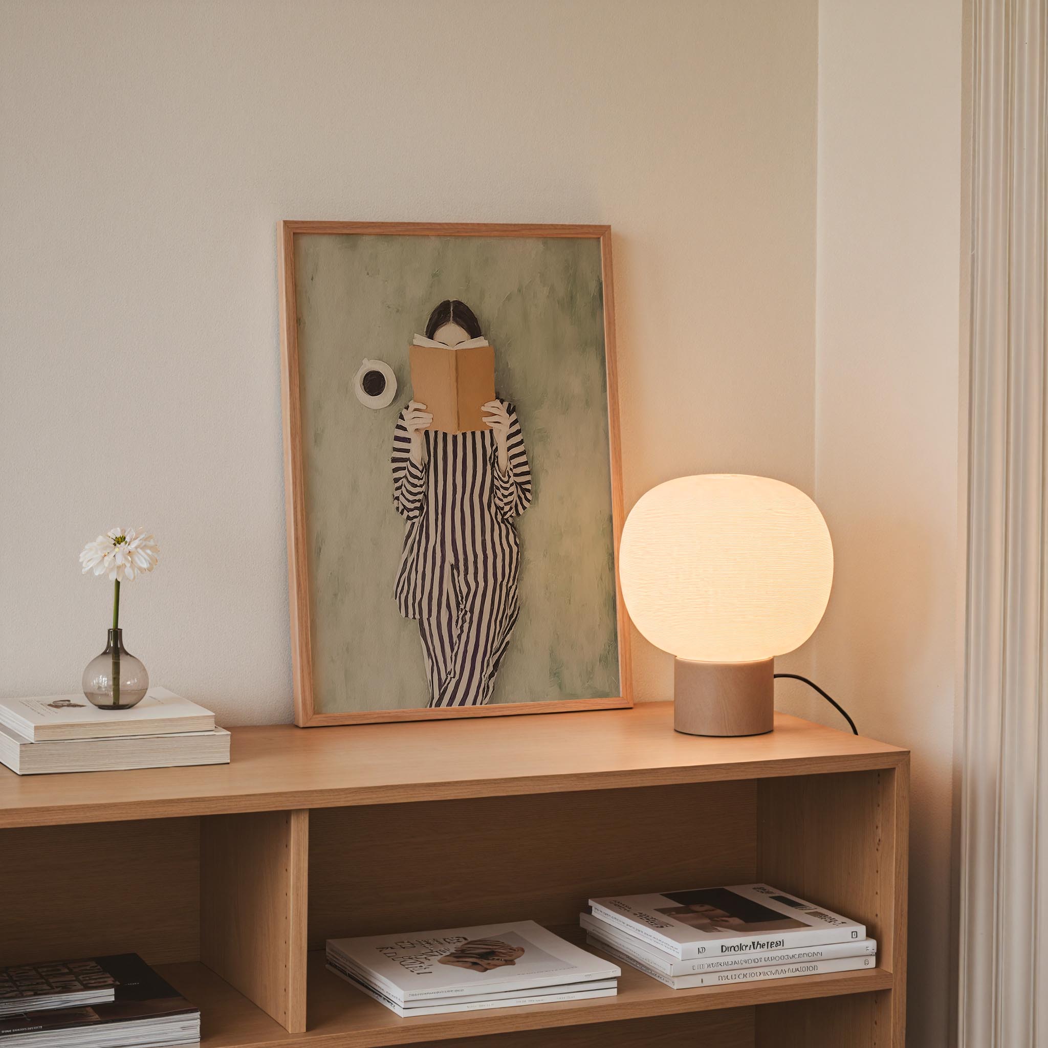 a wooden bookshelf with a framed painting of a woman reading a book, a lamp, and a vase with a single flower.