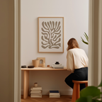 A woman is sitting at a wooden desk, surrounded by various objects, including a plant, books, and a framed abstract art piece on the wall.