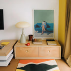a cozy corner of a room with a wooden dresser, a lamp, a framed painting of a woman sitting on a blue couch, and a stack of books.