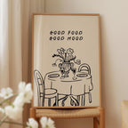 a framed illustration of a table with a vase of flowers, accompanied by the text "GOOD FOOD GOOD MOOD" above it.