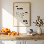 a kitchen counter with a framed poster that reads "Early Morning Coffee Club" above a bowl of oranges and a vase of flowers.